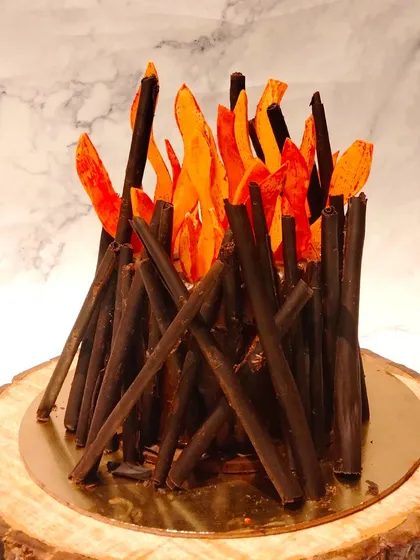A close-up of the Lohri bonfire cake. The chocolate sticks are carefully arranged to resemble logs, and the caramelized sugar flames add a beautiful, warm glow.