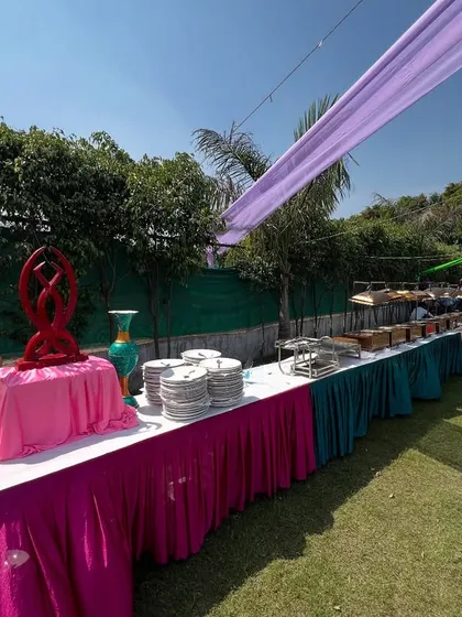 A colorful outdoor buffet setup for a daytime event, with tables draped in bright pink, teal, and magenta fabrics.