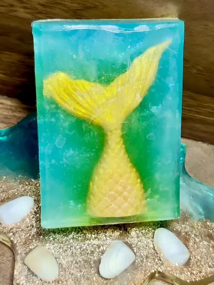 A beautiful mermaid tail soap, displayed on a resin coaster. The clear soap allows the shimmering, colorful tail to be the star of the show.