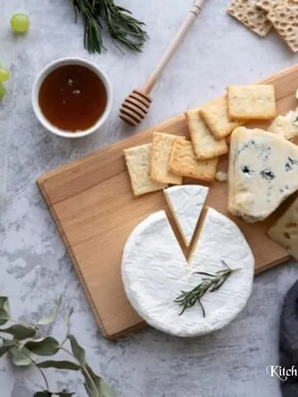 A deconstructed, top-down view of a classic cheese board. This flat-lay shot highlights a wheel of Brie, blue cheese, and hard cheese, accompanied by crackers, grapes, and a bowl of honey.