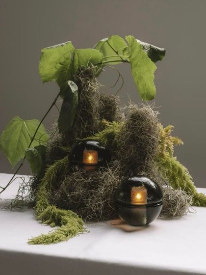 Two smoked grey 14p lamps are artfully placed within a natural arrangement, their dark glass complementing the green foliage.