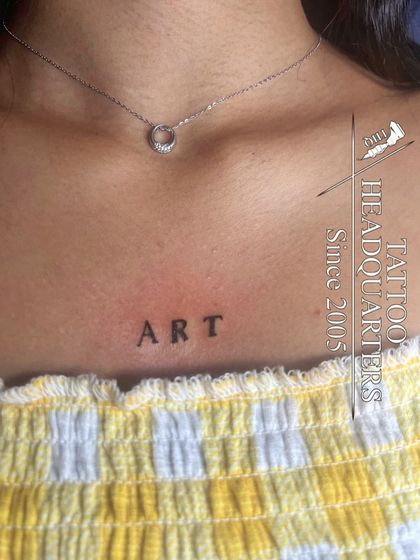 The word "ART" tattooed in a simple, clean font on the collarbone, a perfect piece for an art lover.