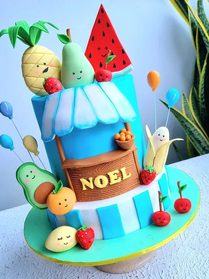 A "tutti frutti" themed cake with a fruit stand and cute, smiling fruit characters all made from fondant. This is a fun design for learning how to model expressive faces.