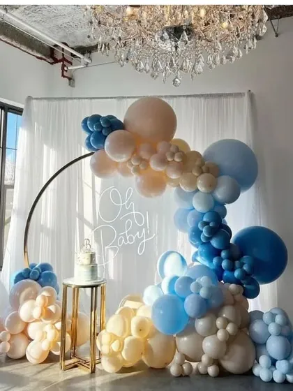 An elegant 'Oh Baby' setup for a baby boy shower. This design uses a circular metal arch and a beautiful cascade of blue, beige, and white balloons, creating a sophisticated backdrop under a crystal chandelier.