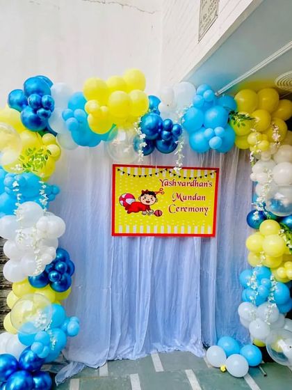 A simple and bright backdrop for "Yashvardhan's Mundan Ceremony." This outdoor setup includes a white curtain, a custom banner, and a colorful balloon arch in yellow, blue, and white.