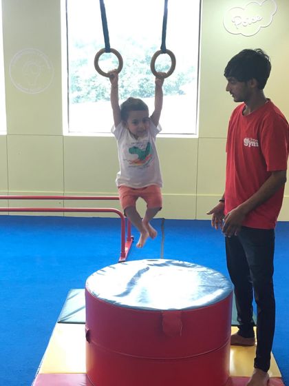 A young boy practices swinging on the gymnastics rings with a coach nearby, developing grip strength and a feel for aerial movement.