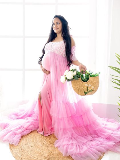 A full-length portrait of a mom-to-be in a flowing pink gown, holding a straw bag with flowers for a fresh, summery feel.