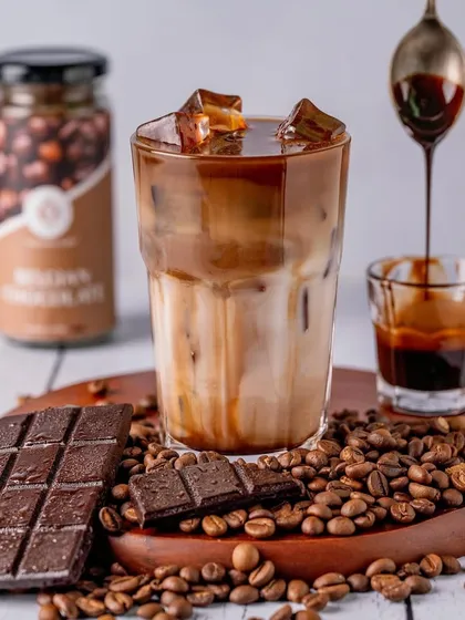 A close-up of a perfectly layered Belgian chocolate iced coffee. The focus on the dripping syrup from the spoon adds a touch of indulgence to this beverage shot.