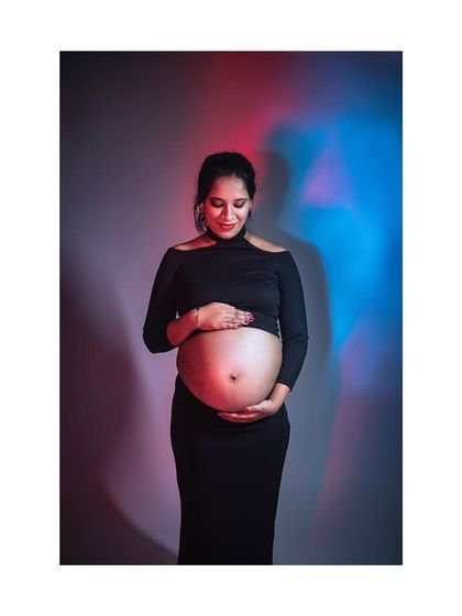 A creative maternity portrait using colored lighting. The mother-to-be looks down at her belly, illuminated by dramatic red and blue lights for a moody and artistic effect.