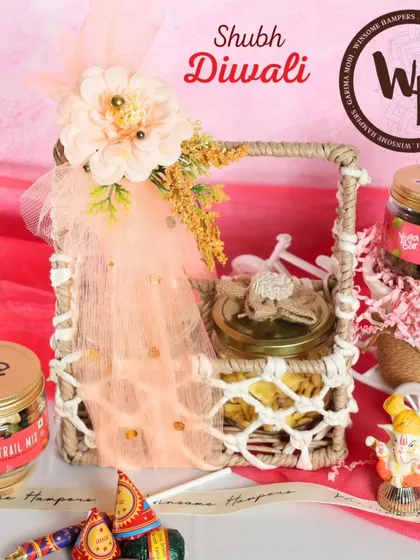 A sweet and simple "Shubh Diwali" hamper. This gift includes jars of trail mix and raisins, a Ganesha figurine, and festive crackers, all beautifully presented in a woven basket with peach-colored netting.