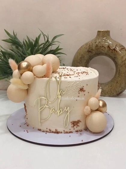 A cutesy and elegant gender-neutral baby shower cake. This design features a clean white finish with beige and gold decorative balls and a lovely "Oh Baby" topper.