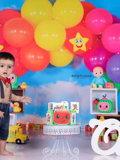 A baby boy stands proudly in his Cocomelon-themed first birthday setup. The bright balloons and character toys create a festive and joyful scene.