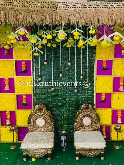 A vibrant setup for an Upanayanam or sacred thread ceremony. The backdrop is a checkerboard of yellow marigolds and pink fabric, with hanging bells and two traditional chairs for the ceremony.