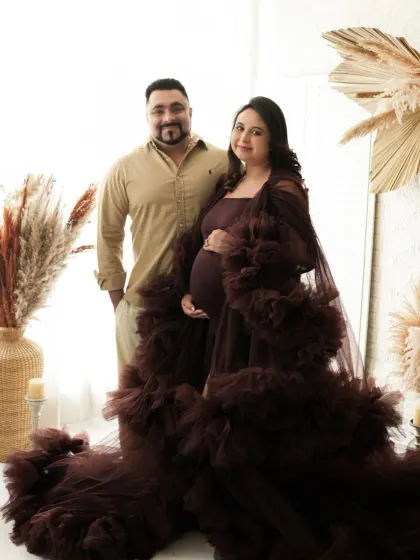 A classic standing portrait of an expecting couple. The rich brown gown and neutral studio background create a sophisticated and timeless image.