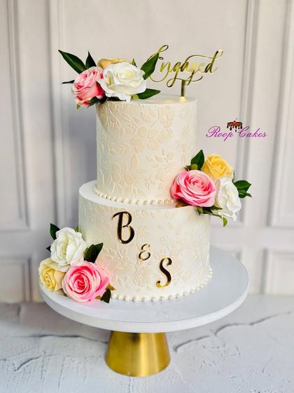 This classic two-tier engagement cake features a subtle floral stencil pattern on white whipped cream. It's adorned with beautiful pink, yellow, and white roses and a golden 'Engaged' topper for a touch of elegance.