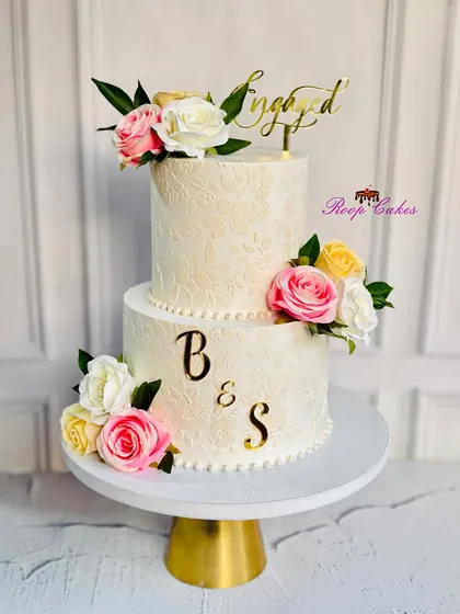 Celebrating love with sweetness and style. This two-tier engagement cake features a subtle white-on-white stencil design, adorned with pink, yellow, and white roses and personalized with the couple's initials.