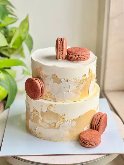 This modern two-tier cake features a textured white buttercream base with abstract gold leaf detailing. The design is topped with rich, chocolate-colored macarons for a contemporary finish.