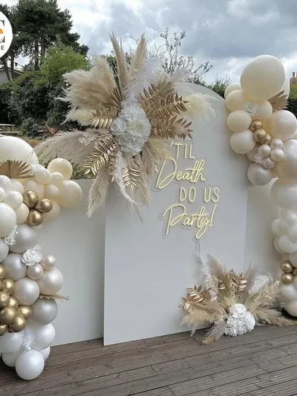A stylish and modern party backdrop with white and gold balloon arches and pampas grass, perfect for a bridal shower or engagement party.