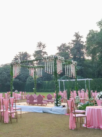 A wide shot of the garden wedding setup, showing the pink-themed guest seating arranged around the beautiful floral mandap. The open-air setting and coordinated colors create a harmonious and romantic atmosphere.
