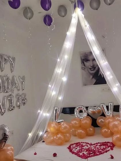 A romantic bedroom decoration for a birthday, featuring a canopy with fairy lights, "LOVE" balloons, and a heart made of rose petals on the bed.