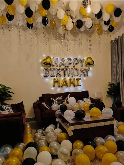 A birthday living room decoration in black, white, and gold. The setup includes personalized foil letters, balloons on the ceiling and floor, and fairy lights.
