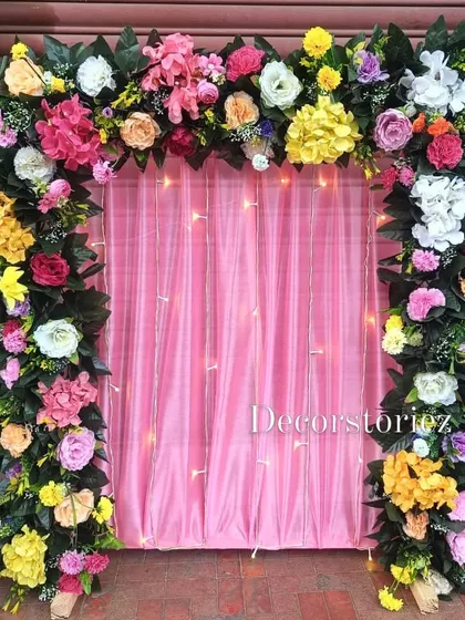 This is one of our ready-to-install backdrops, featuring a lush arch of multi-colored artificial flowers over a pink satin curtain with delicate fairy lights. It's a versatile setup suitable for home pujas, birthdays, or festival celebrations.
