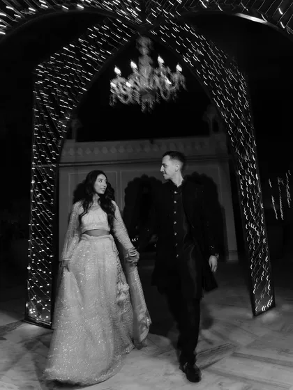 A candid black-and-white shot of the couple walking hand-in-hand under a beautifully lit archway, capturing a fairytale-like moment from their reception.