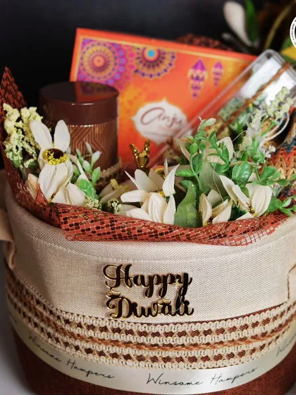 A close-up of my "Happy Diwali" fabric basket hamper. This view shows the arrangement of a scented candle, a box of treats from the brand Anjeer, and delicate white flowers, creating a warm and festive feel.