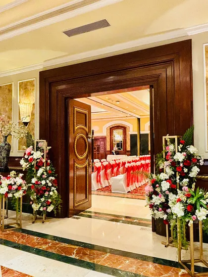 The view from the entrance looking into the main event hall. The doorway is flanked by tall floral arrangements in red and white, giving guests a tantalizing glimpse of the celebration inside.