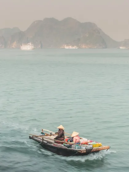 A wide shot showing a small vendor boat navigating the vast, misty landscape of Halong Bay.