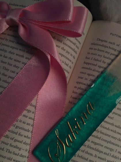 A close-up of the 'Sakina' bookmark, styled with a pink ribbon. The details of the gold name and the teal resin are clearly visible.