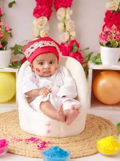 This baby looks directly at the camera, showing off his festive red headwear and the colorful Holi setup.