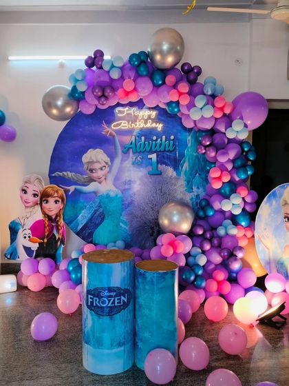 A magical Frozen theme for a first birthday. The decoration includes a custom circular backdrop with Elsa, a beautiful balloon garland in shades of purple, blue, and pink, and a marquee number '1'.