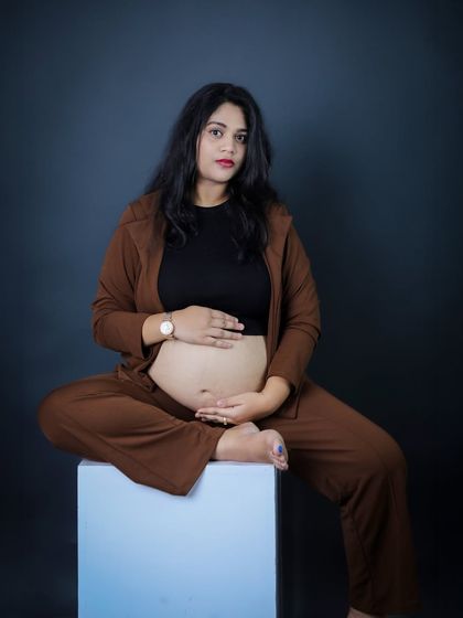A powerful and modern studio portrait. The mother-to-be sits confidently in a stylish brown suit, embracing her baby bump against a dark, moody background.