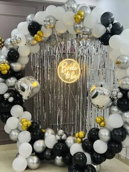 A chic and glamorous "Bride to be" party setup with a black, white, and silver theme. A full circular balloon arch surrounds a shimmering silver tinsel backdrop, with a neon sign and disco ball accents for a fun, party ready photo corner.