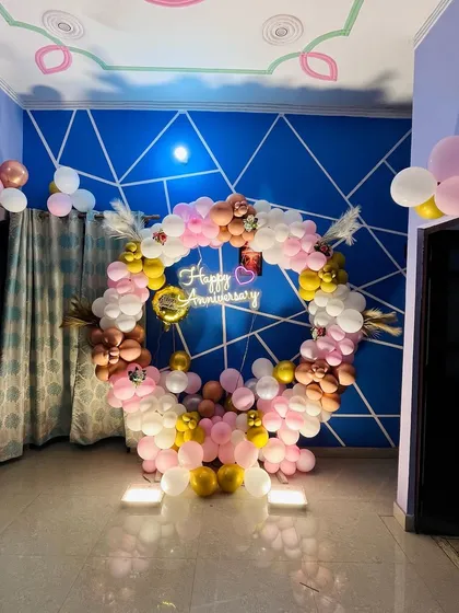 A full circular balloon arrangement in pink, white, and gold for an anniversary celebration at home, complete with a neon sign and feather accents.
