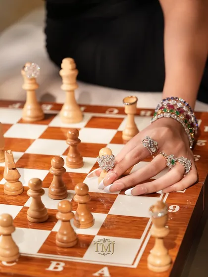 A close-up of a hand adorned with statement rings making a move on a chessboard, where each chess piece is also topped with a diamond ring.