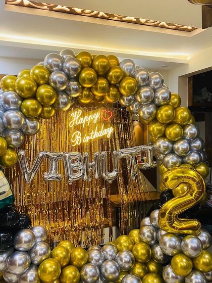 A grand 25th birthday celebration with a gold shimmer wall, a massive gold and silver balloon arrangement, and a champagne bottle prop.