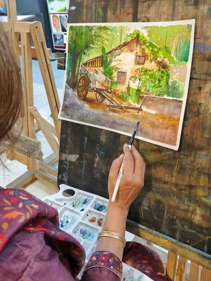 An artist at her easel, working on a watercolor painting of a village scene. This shot highlights the tools of the trade, including the palette with its pools of color.