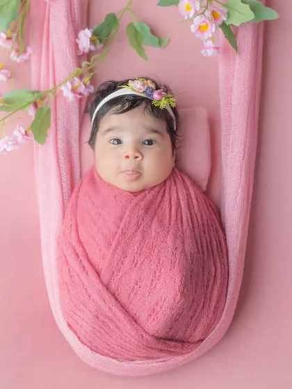 An adorable awake newborn, snugly wrapped and hanging in a soft pink sling surrounded by blossoms.