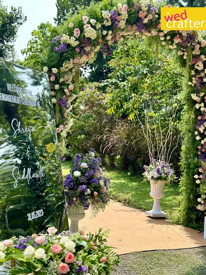 A stunning floral arch covered in lilac, lavender, and white flowers welcomes guests to an elegant garden engagement ceremony.