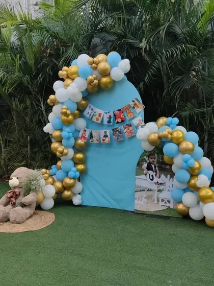 A whimsical outdoor photo booth for a first birthday, with a teddy bear prop and a lovely balloon arch against a blue backdrop.
