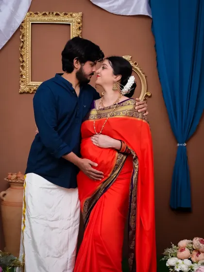 A loving portrait of a couple in a traditional setting. The mom-to-be is radiant in a red and purple saree, while her partner complements her in a dhoti. This setup is designed to celebrate your journey with cultural richness and elegance.