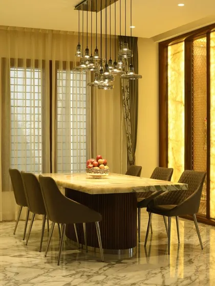 This elegant dining setup features a backlit onyx wall that casts a warm, inviting glow. The marble-topped table and plush dining chairs are illuminated by a modern cluster chandelier, creating a luxurious atmosphere for meals.