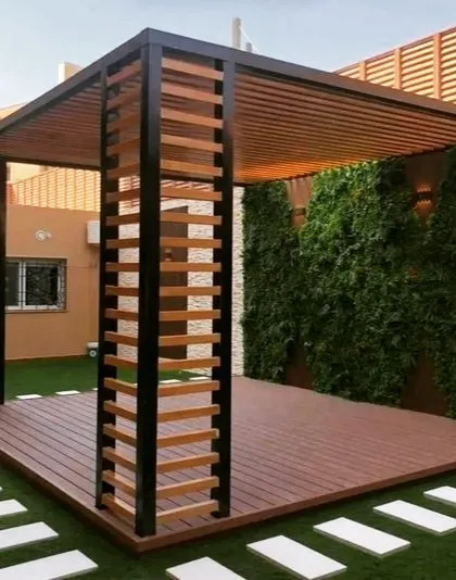 A unique pergola design with decorative wooden slats on the support columns. The structure is set on a composite wood deck and features an artificial green wall backdrop.