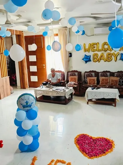 A simple and sweet living room decoration for a baby boy's welcome. The setup includes balloons on the ceiling, a "WELCOME BABY" foil display, and a heart shape made of rose petals on the floor.