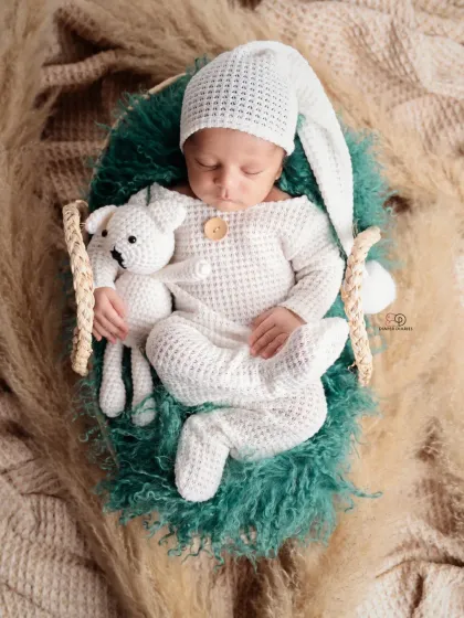 A sleeping newborn in a cozy, textured white romper and matching hat, holding a small knitted toy. This boho-inspired shot uses natural textures like pampas grass for a warm, earthy feel.