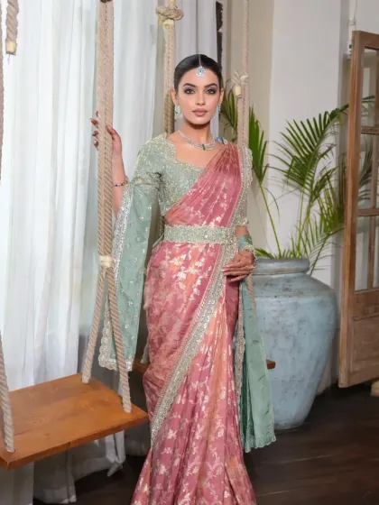A full-length portrait from a designer shoot featuring a South Indian bridal look. The hair is styled in a neat bun and a long, floral gajara braid, complementing the elegant saree.