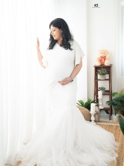 A serene moment by the window. The soft, natural light beautifully highlights the mom-to-be's silhouette and the delicate details of her white gown.