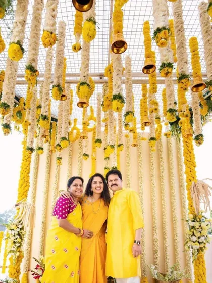A happy family posing in front of a haldi backdrop I designed. The ceiling is covered with hanging strings of white and yellow flowers with small bells, creating a beautiful and festive atmosphere.